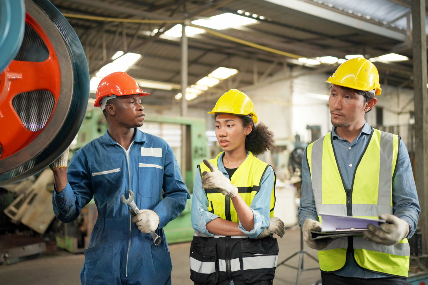 Three Diverse Multicultural Heavy Industry Engineers and Workers in Uniform at Steel Factory. Équipe d’ingénieurs et d’opérateurs en discussion devant une machine dans un atelier industriel, portant des casques et gilets de sécurité, illustrant la performance et la collaboration en production.